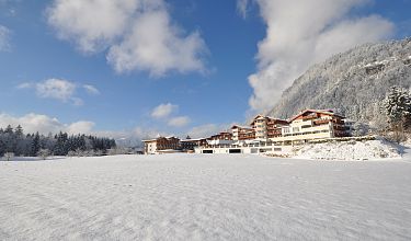 Winterlandschaft mit schneebedecktem Feld, im Hintergrund ein großes Gebäude und bewaldete Berge unter einem strahlend blauen Himmel.