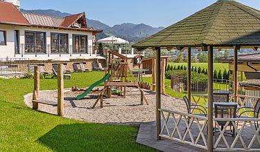 Spielplatz mit Holzrutsche und Turm auf grünem Rasen. Im Hintergrund Berge und ein Gebäude mit Terrasse.