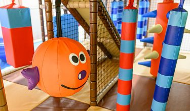 Ein farbenfroher Indoor-Spielplatz mit großen Schaumstoffspielzeugen, darunter ein orangefarbener Ball mit Gesicht und hängende Zylinder.