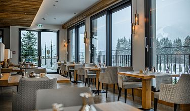 Modernes Restaurant mit großen Fenstern, durch die verschneite Landschaft sichtbar ist. Holzverkleidung und graue Polstermöbel dominieren das Interieur.