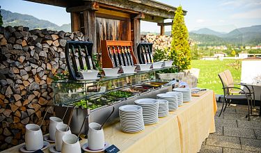 Ein Buffet mit Tellern und Besteck vor einer Berglandschaft auf einer Terrasse. Der Himmel ist klar, mit Bergen und Holzstapeln im Hintergrund.