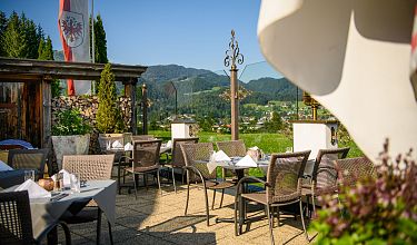 Gemütliche Terrasse eines Restaurants mit gedeckten Tischen, umgeben von grüner Landschaft und Bergen im Hintergrund.