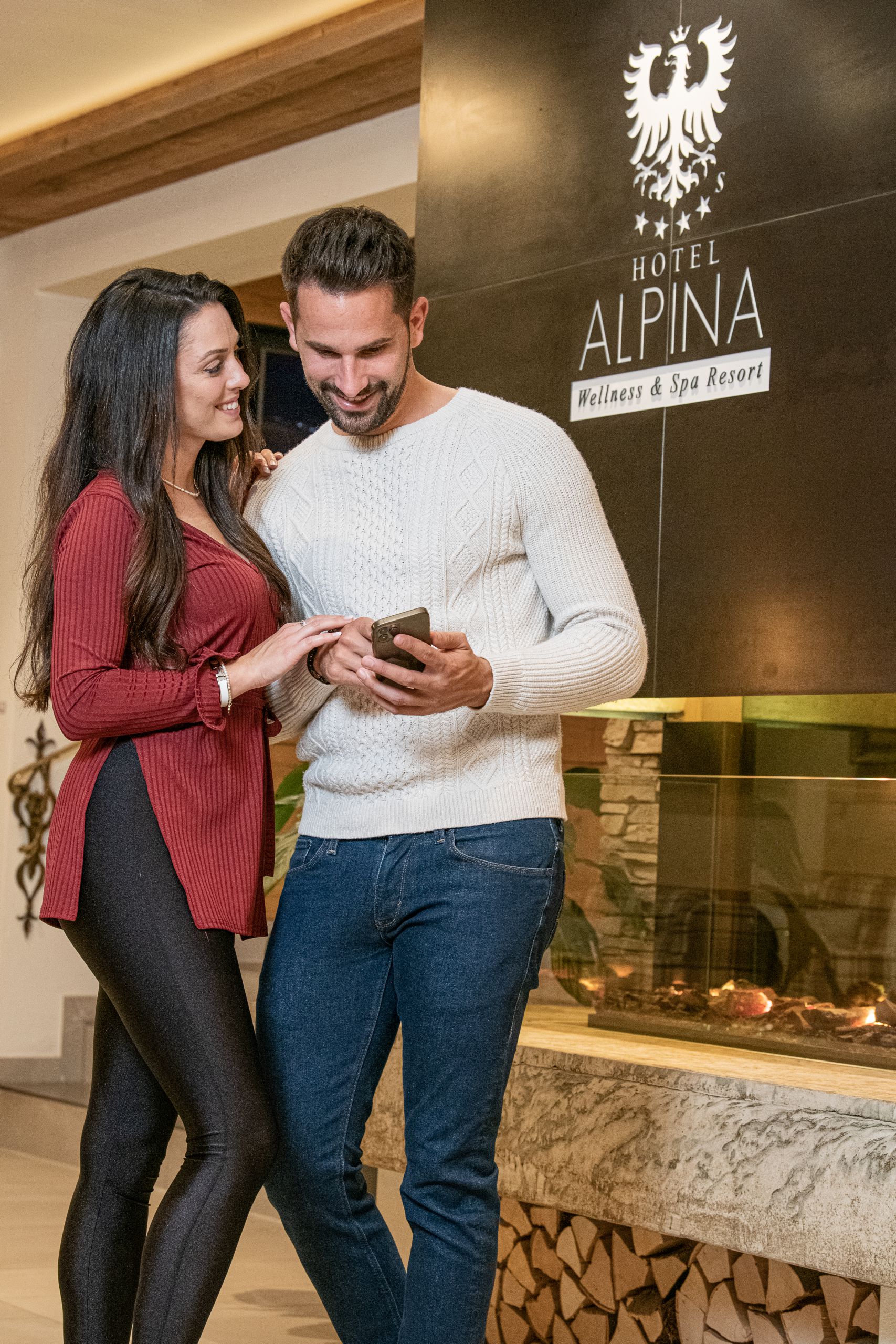 Lächelndes Paar mit Handy; sie tragen Freizeitkleidung. Im Hintergrund sieht man ein Schild für ein Wellness & Spa Resort.