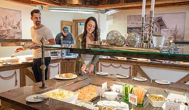 Menschen bedienen sich an einem Buffet mit verschiedenen Speisen. Der Raum ist rustikal dekoriert mit Holzverkleidung und Kerzen.