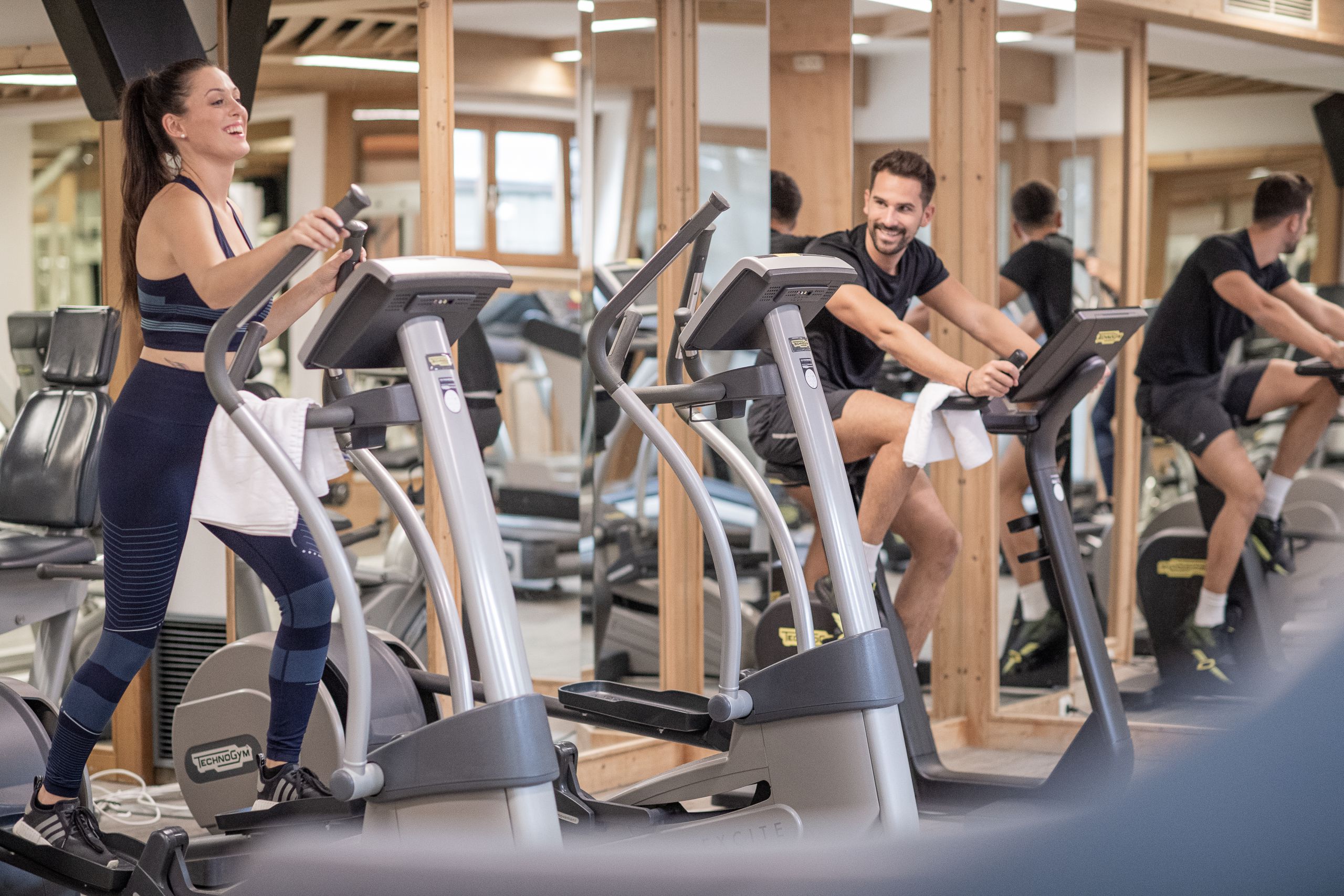 Menschen trainieren in einem Fitnessstudio. Eine Frau läuft auf einem Laufband, während ein Mann auf einem anderen Gerät lächelt.